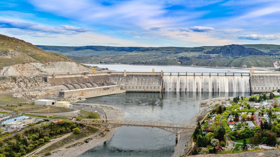 A large dam with a bridge crossing a river, surrounded by hills and a small town.