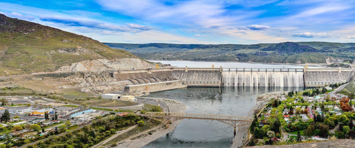 A large dam with a bridge crossing a river, surrounded by hills and a small town.