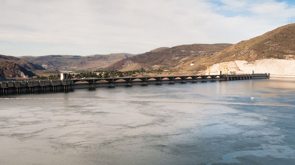 A dam with a bridge over a calm lake, surrounded by hills and mountains.