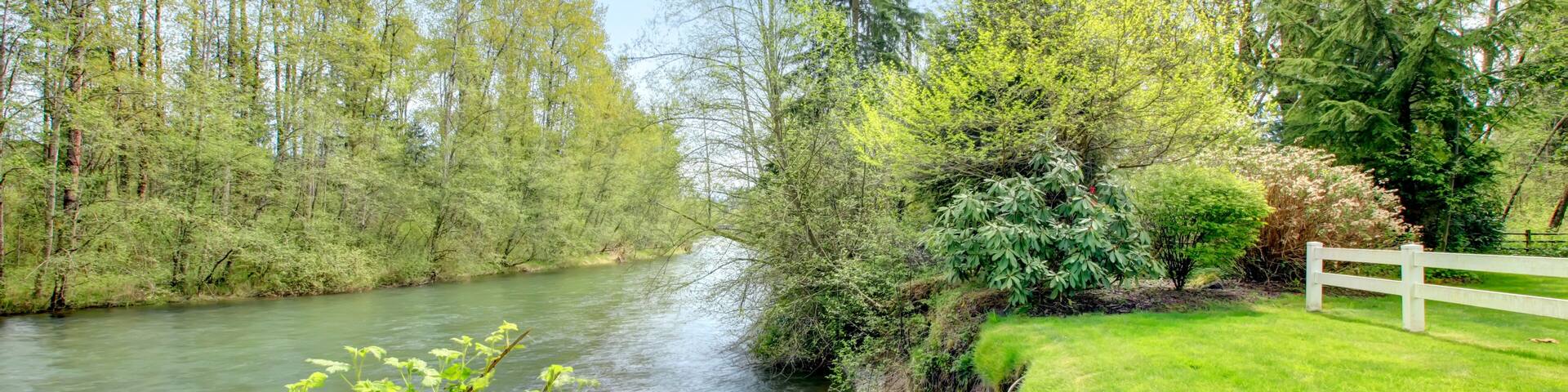 A river flowing through a lush green landscape with trees and a white fence along the riverbank.