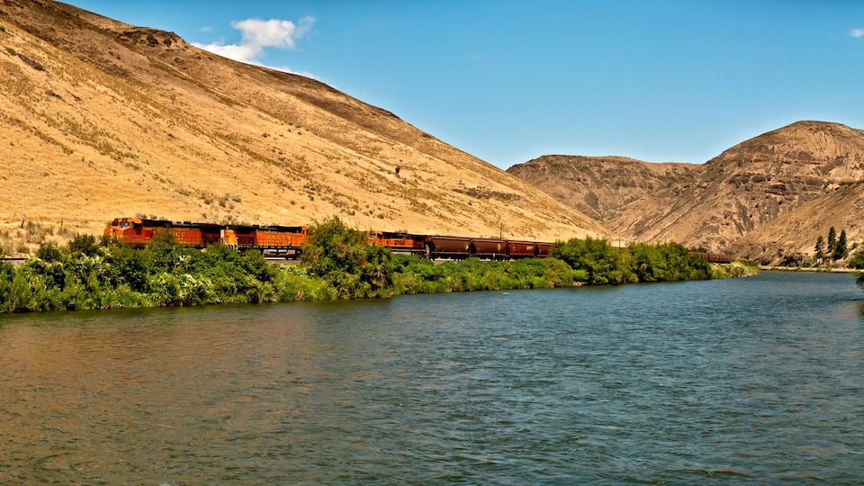 A river with a train on a bridge, surrounded by mountains.
