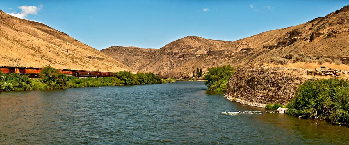 A river with a train on a bridge, surrounded by mountains.