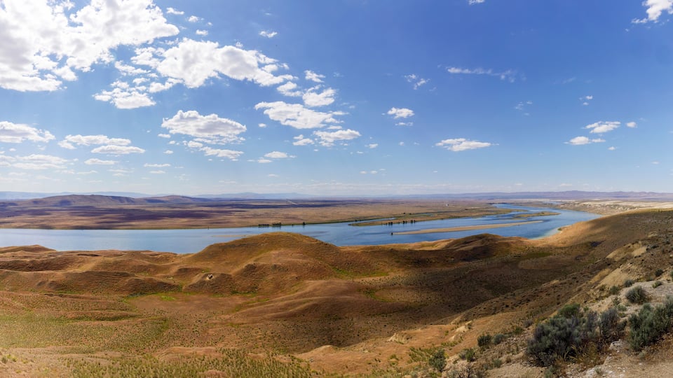 A vast landscape with rolling hills, a river, and distant mountains under a clear blue sky.