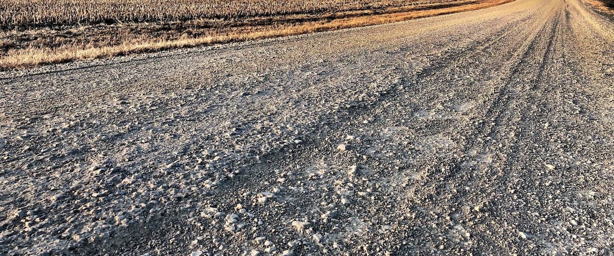 A gravel road in a rural area with fields and farm buildings.