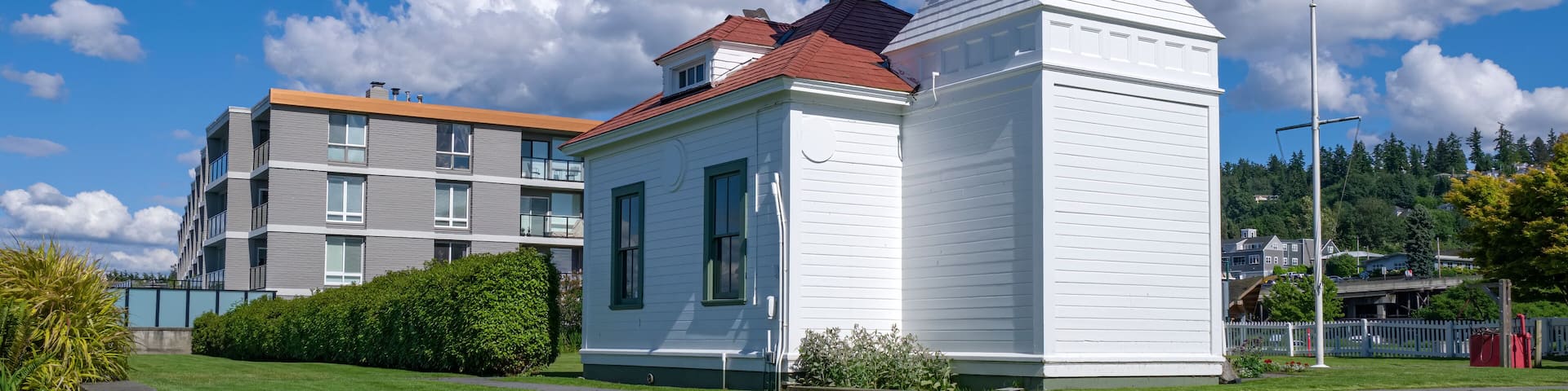 A white lighthouse with a red roof, surrounded by a well-maintained lawn and a modern building in the background.