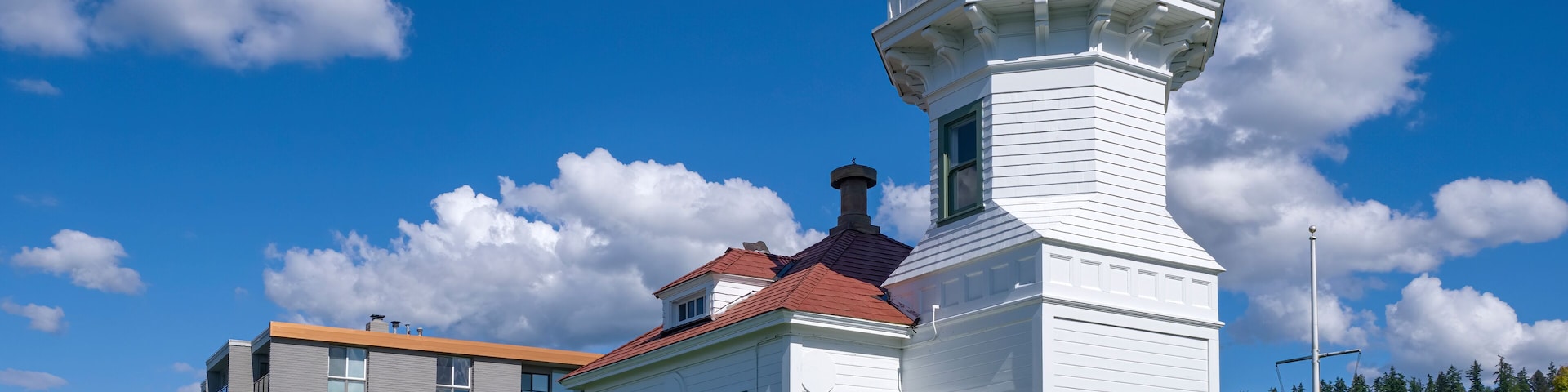 Um farol branco com telhado vermelho, cercado por um gramado bem cuidado e um edifício moderno ao fundo.
