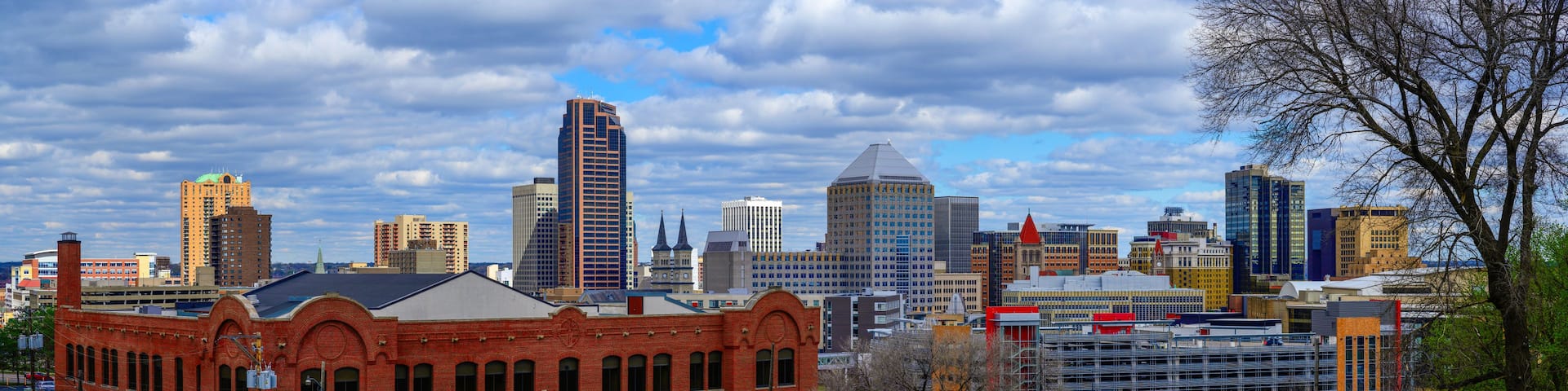 A city skyline with a mix of modern and historic buildings, a prominent red brick building, and a clear blue sky with clouds.