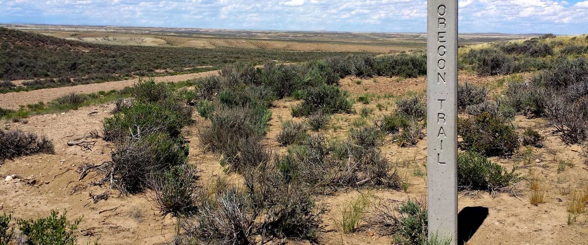A sign reading "OREGON TRAIL" in a desert landscape with sparse vegetation and a clear sky.