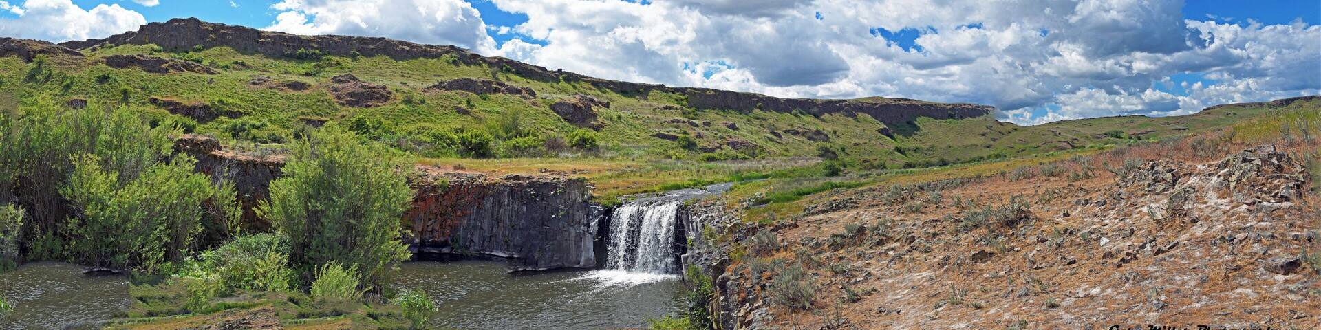A waterfall in a green valley with a rocky cliff and a clear blue sky.