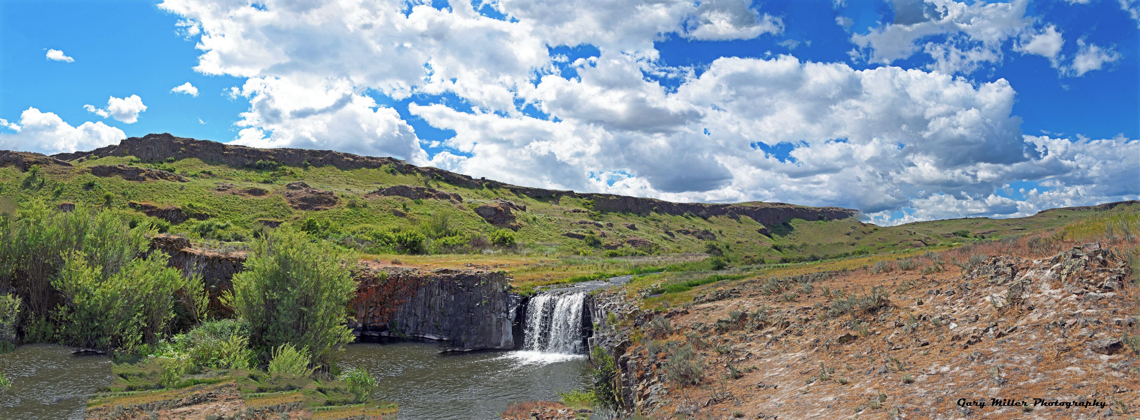 A waterfall in a green valley with a rocky cliff and a clear blue sky.