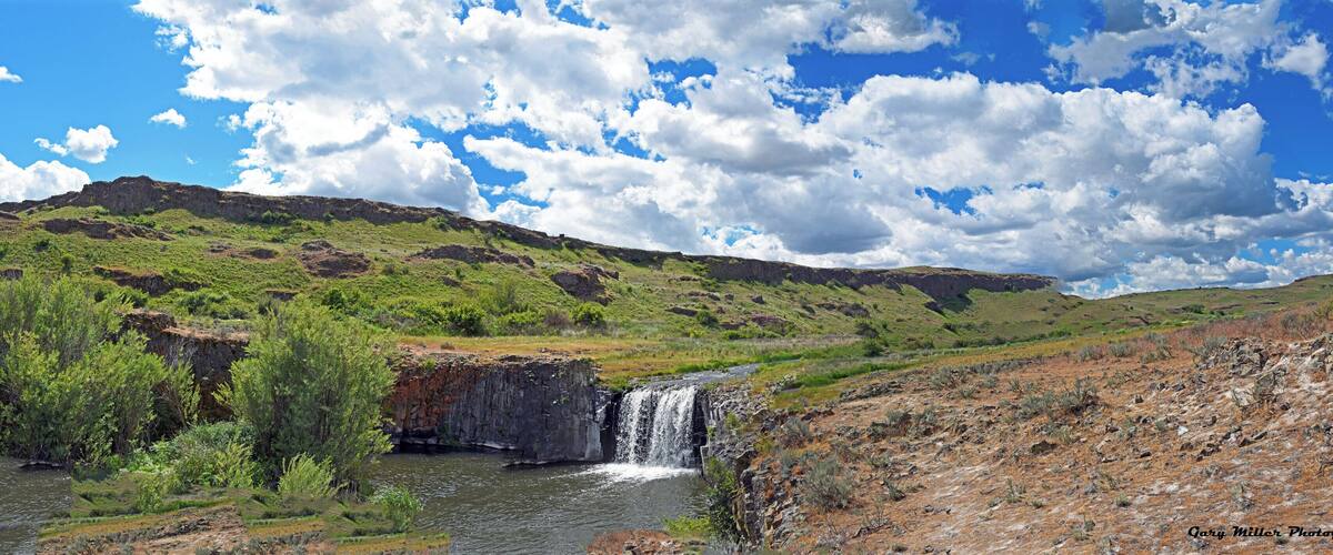A waterfall in a green valley with a rocky cliff and a clear blue sky.