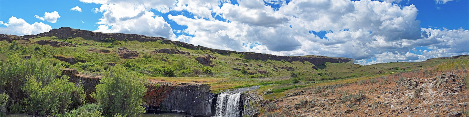 A waterfall in a green valley with a rocky cliff and a clear blue sky.