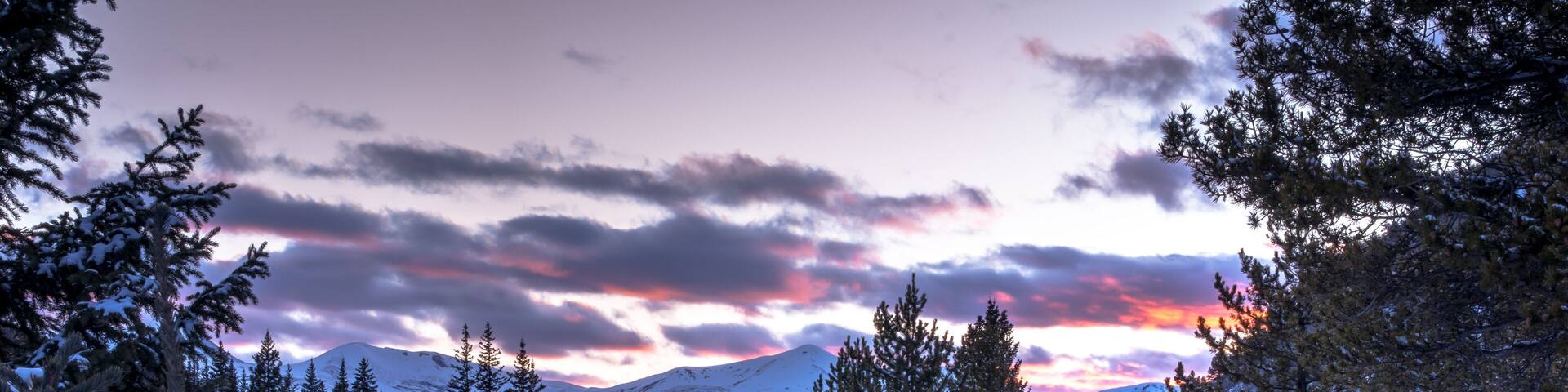 A snowy mountain landscape with evergreen trees and a sunset sky.