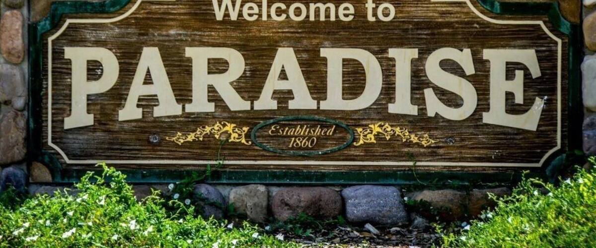 A wooden sign with the text "Welcome to PARADISE Established 1860" surrounded by greenery.