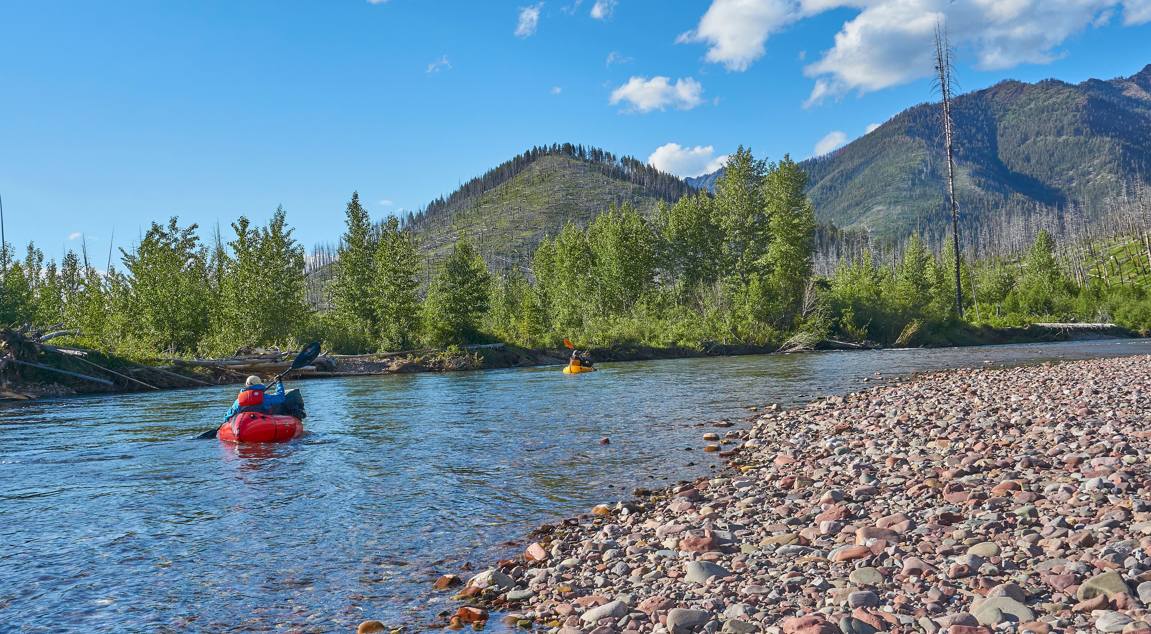 A river with kayakers, surrounded by mountains and a pebble beach.