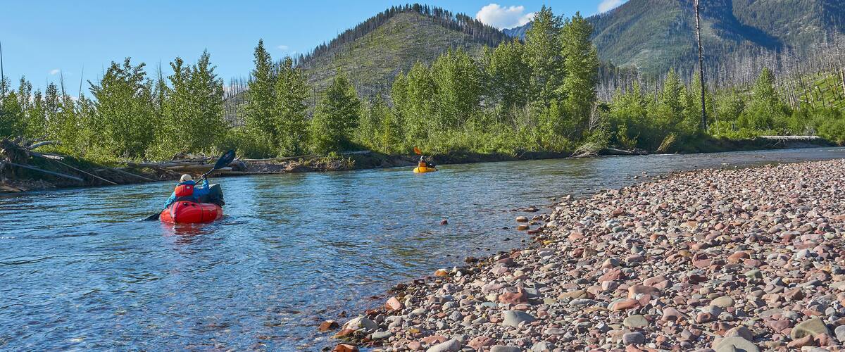 A river with kayakers, surrounded by mountains and a pebble beach.