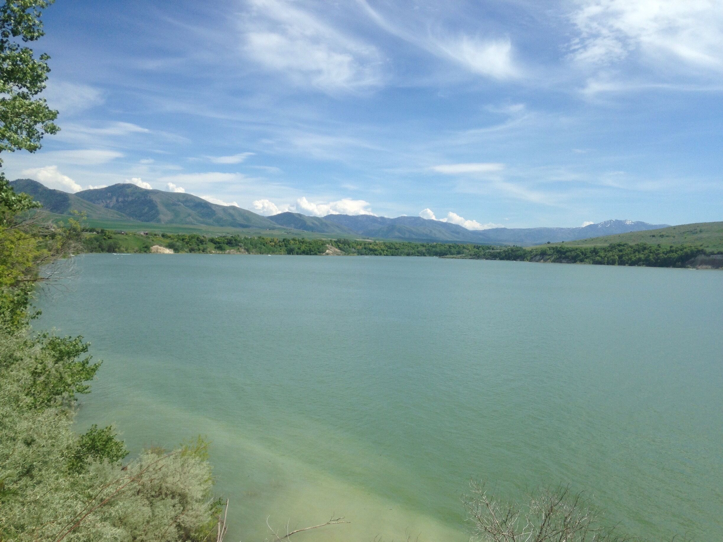 A calm lake surrounded by green hills and mountains under a partly cloudy sky.