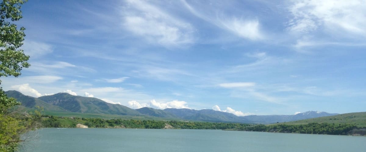 A calm lake surrounded by green hills and mountains under a partly cloudy sky.