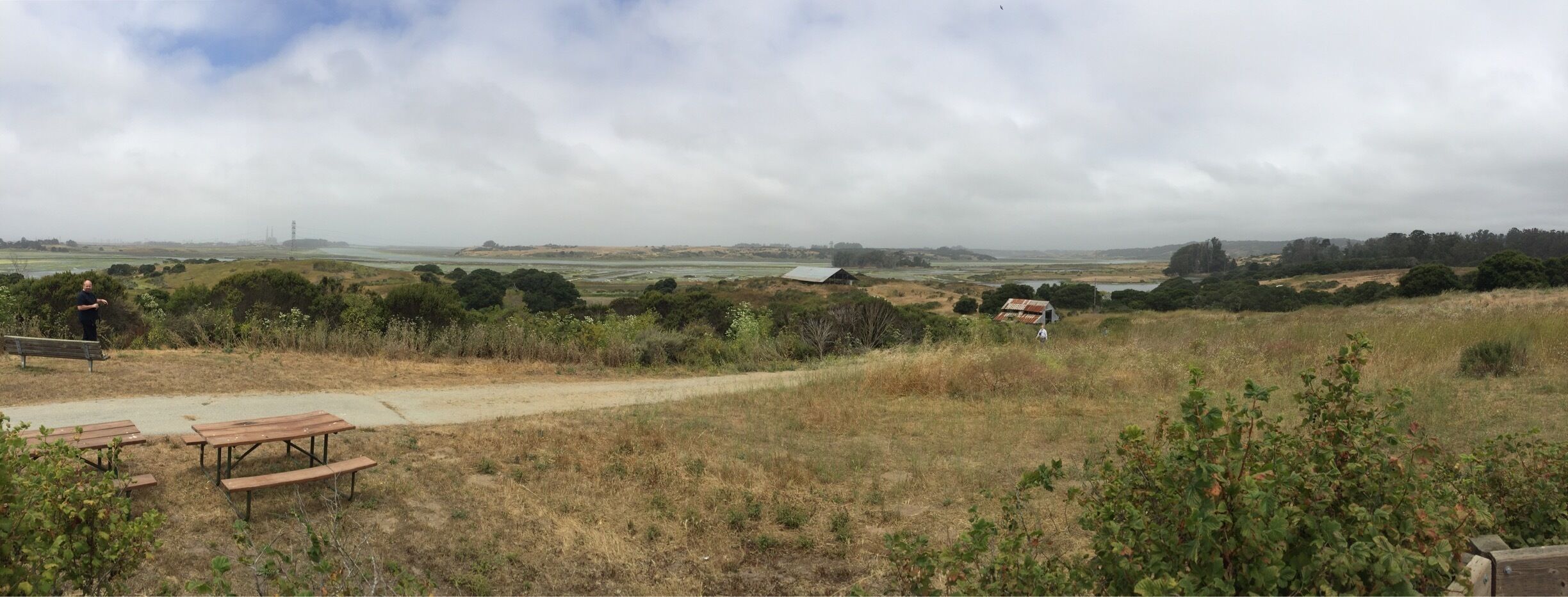 A rural landscape with a dirt path, picnic tables, and a small building.