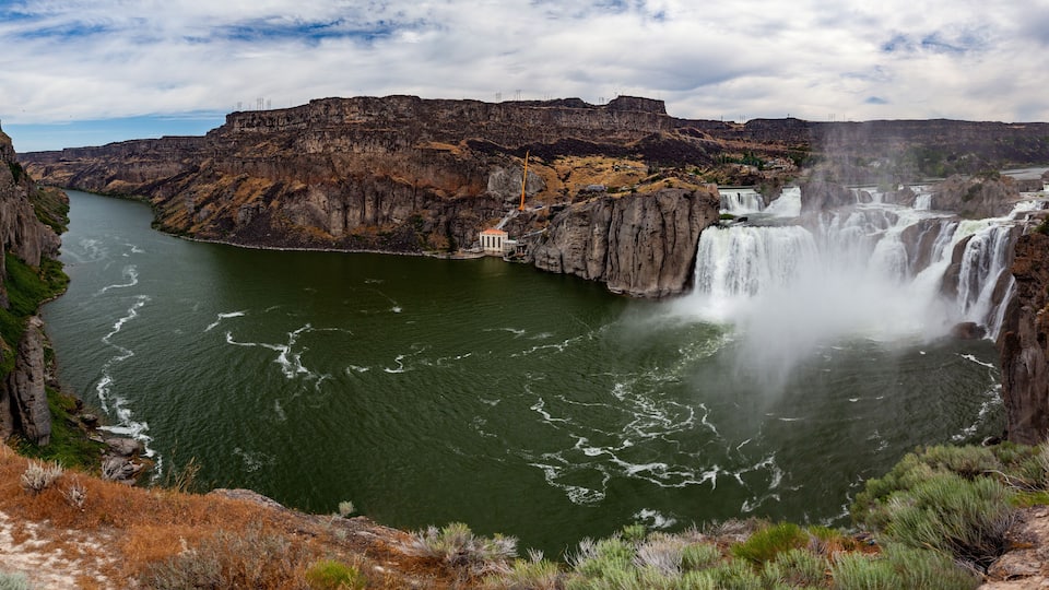 A large waterfall cascading down a steep cliff into a river surrounded by rocky terrain and sparse vegetation.