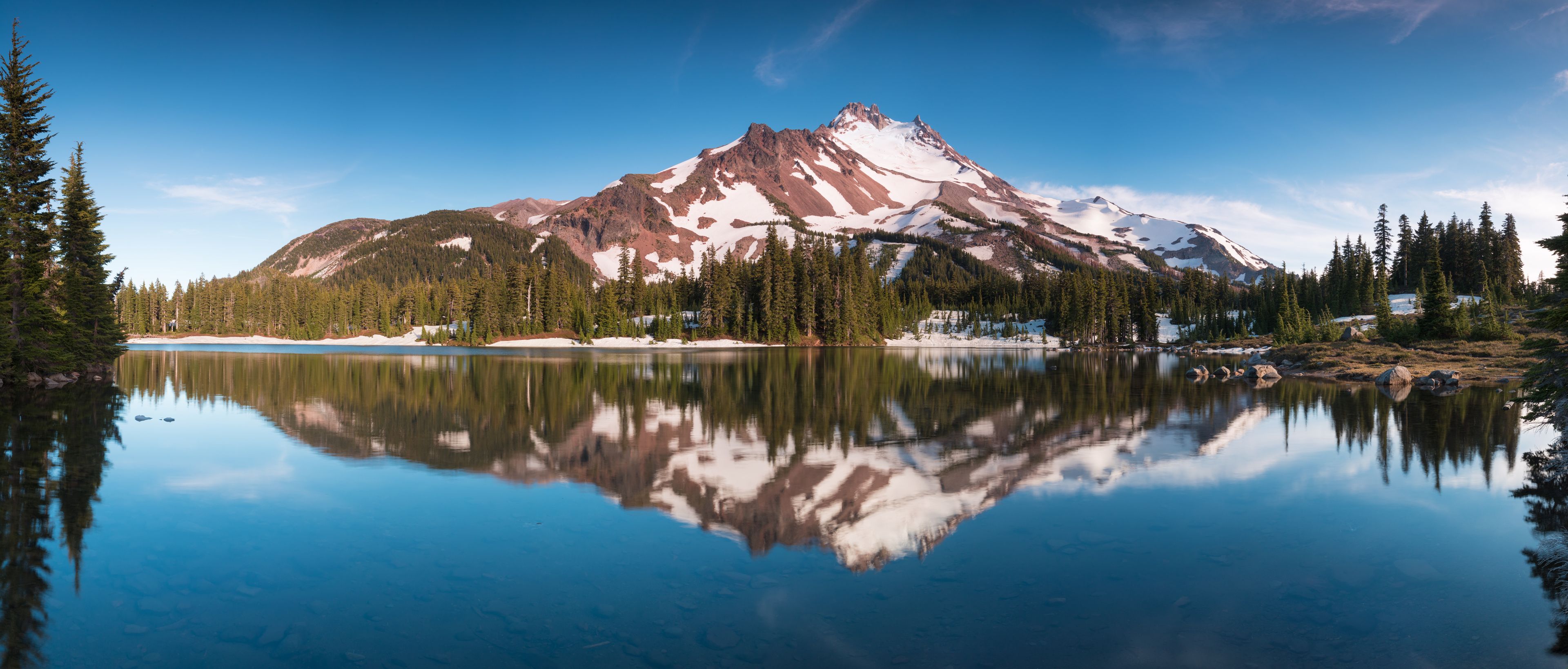 A mountain range with snow-capped peaks, a calm lake reflecting the scenery, and dense coniferous forests.