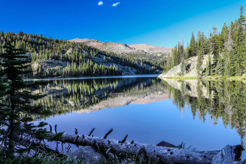 A calm lake surrounded by dense forests and mountains under a clear blue sky.