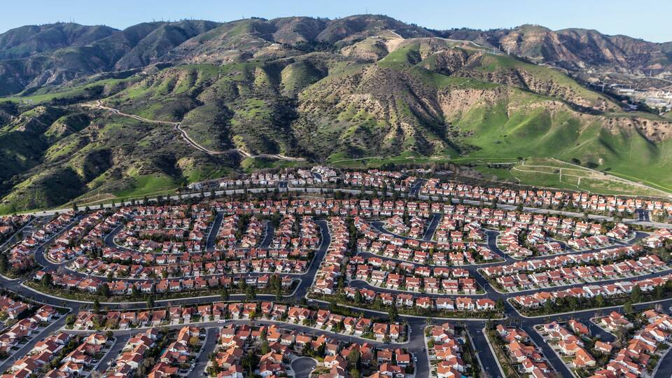 A residential area with winding roads and houses, surrounded by green hills and mountains.