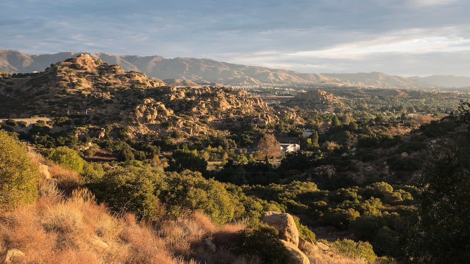 A mountainous landscape with a valley, scattered trees, and distant buildings.