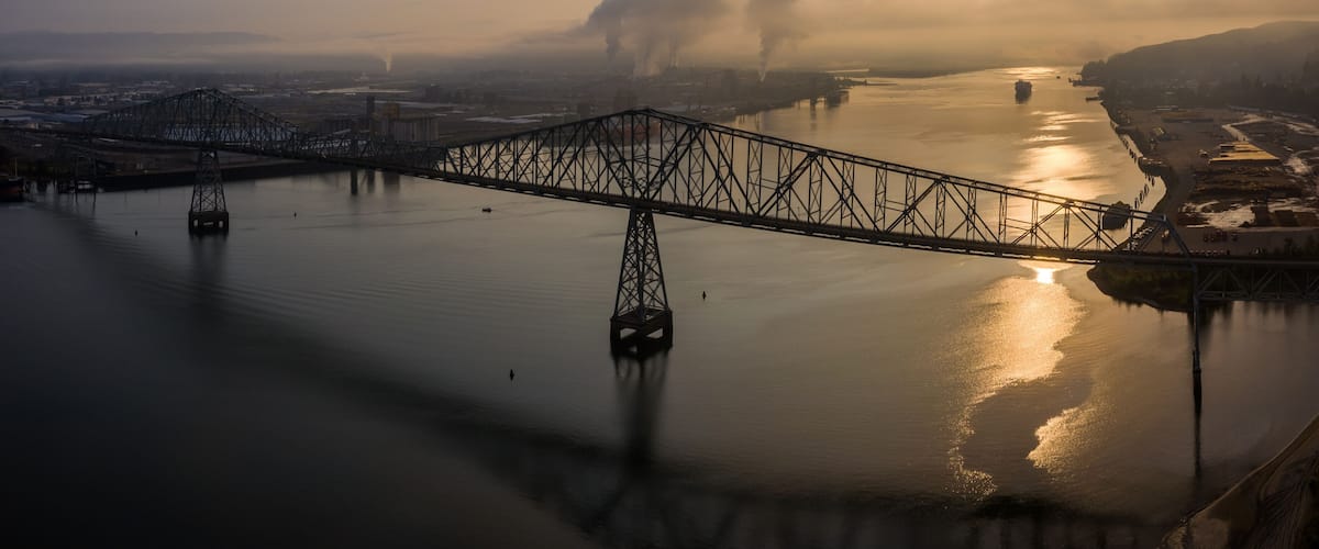 A steel bridge spanning a calm river with industrial buildings and smoke in the background.