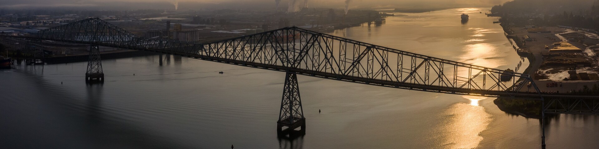 A steel bridge spanning a calm river with industrial buildings and smoke in the background.