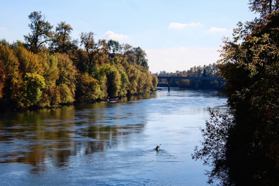 A calm river with a kayaker, surrounded by autumn foliage and a bridge in the background.