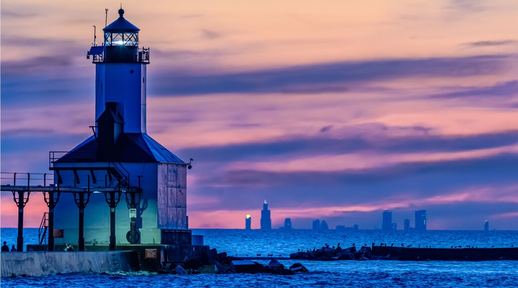 A lighthouse at sunset with a city skyline in the background.