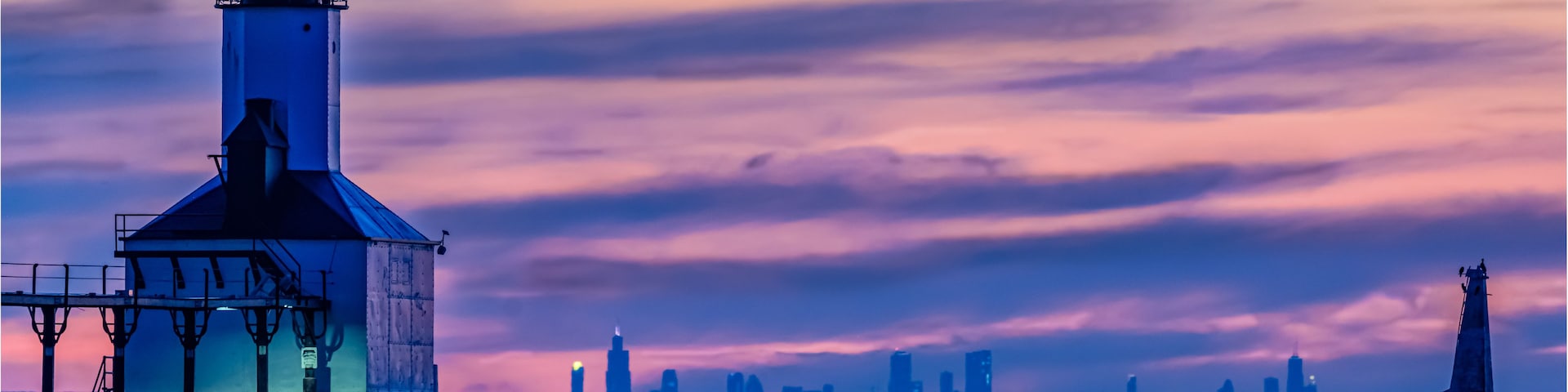 Un faro al atardecer con el perfil urbano de una ciudad al fondo.