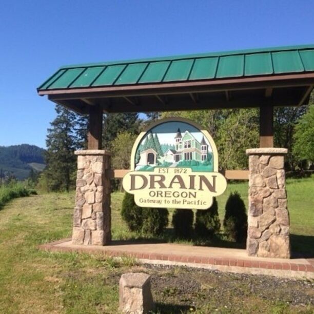 A sign for Drain, Oregon, established in 1872, with a green roof and stone pillars.