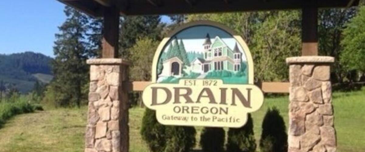 A sign for Drain, Oregon, established in 1872, with a green roof and stone pillars.