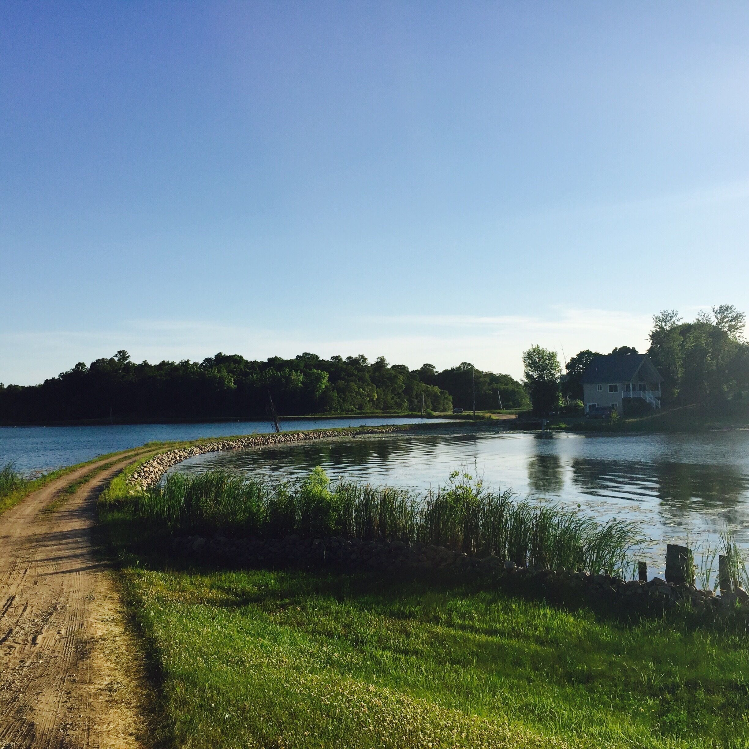 A dirt path beside a calm lake with a stone wall and grassy banks.