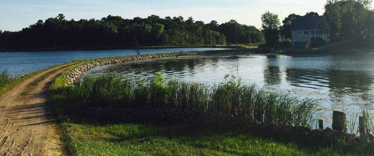 A dirt path beside a calm lake with a stone wall and grassy banks.