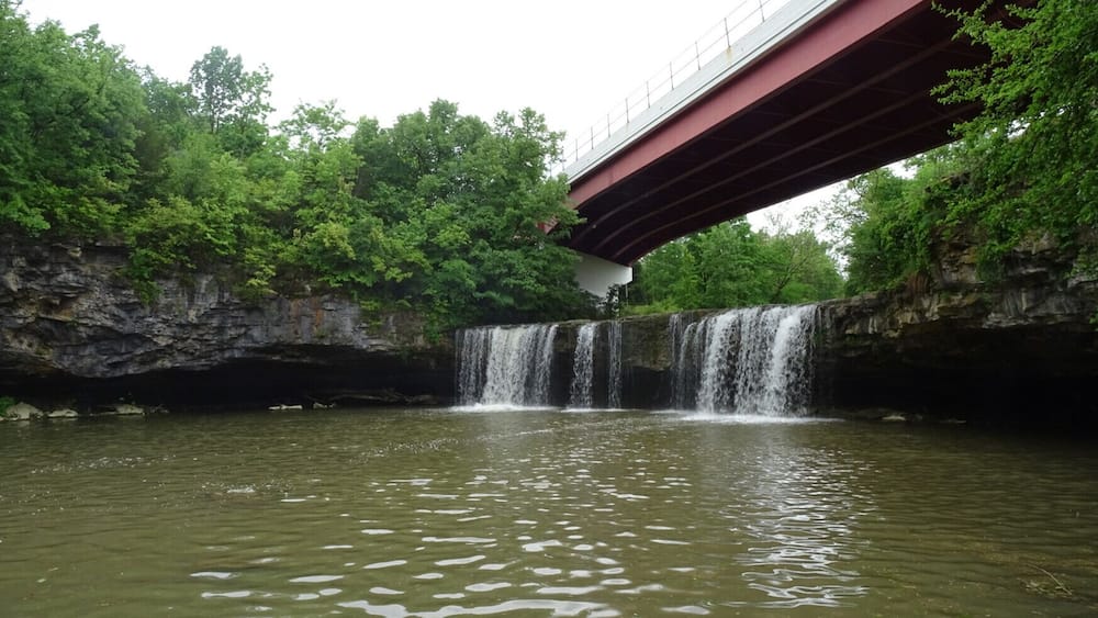 A waterfall under a bridge with lush greenery around.