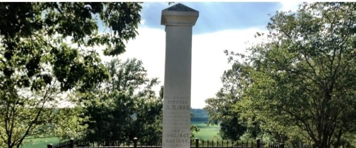 A tall monument with engraved text, surrounded by trees and a fence.