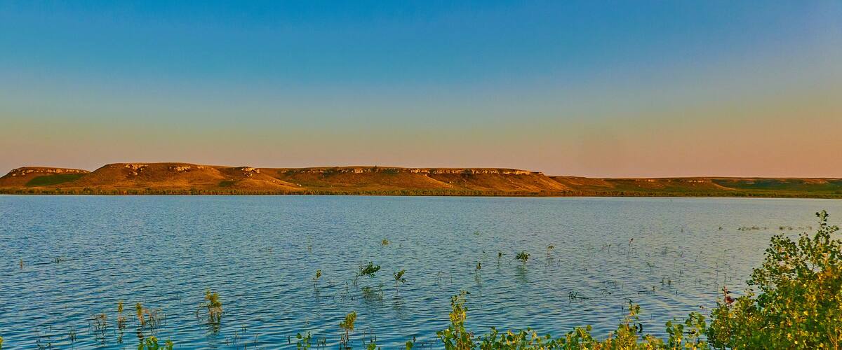 A calm lake with a distant hill range and sparse vegetation.