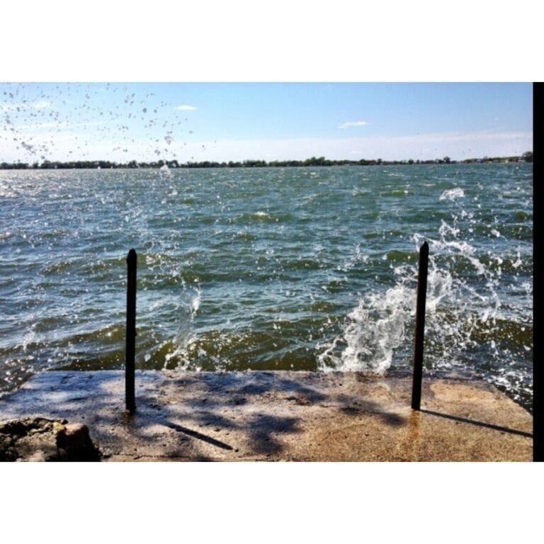 A concrete pier with water splashing against it, a clear sky, and a distant shoreline.