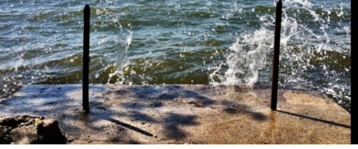A concrete pier with water splashing against it, a clear sky, and a distant shoreline.