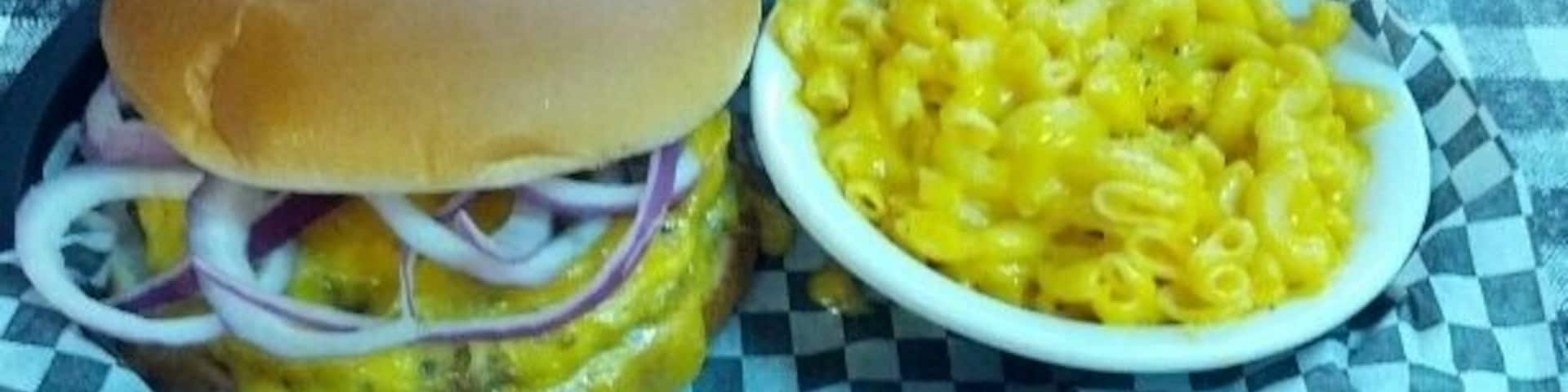 A burger with a bun, cheese, and onions, and a bowl of macaroni and cheese on a checkered tablecloth.