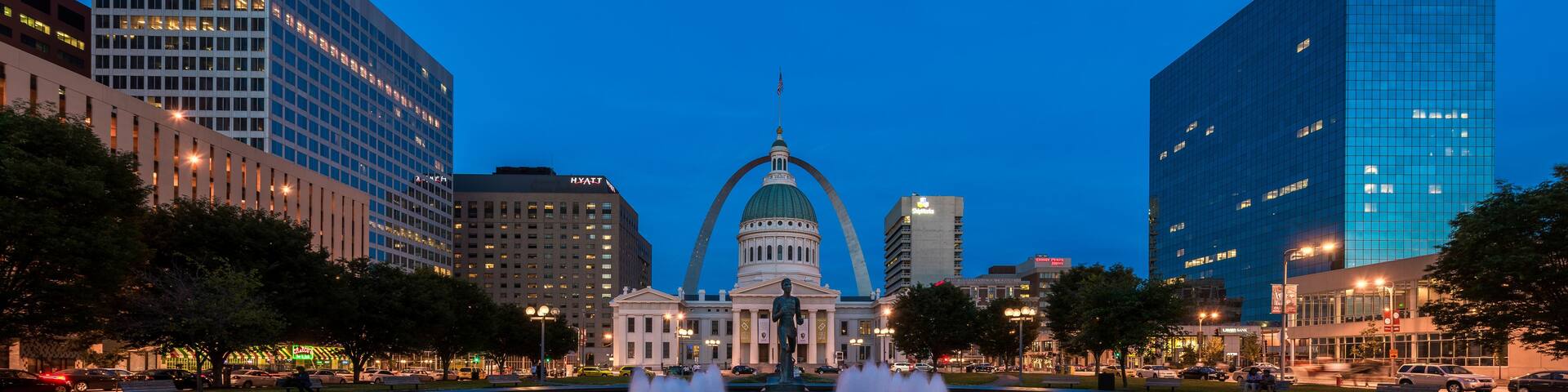 A cityscape at dusk with a prominent building with a dome and an arch structure in the center, surrounded by modern high-rise buildings and a circular fountain.