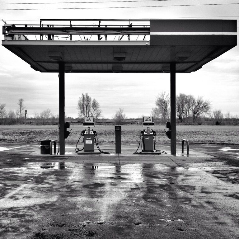A gas station with two pumps and a canopy, surrounded by a rural landscape.