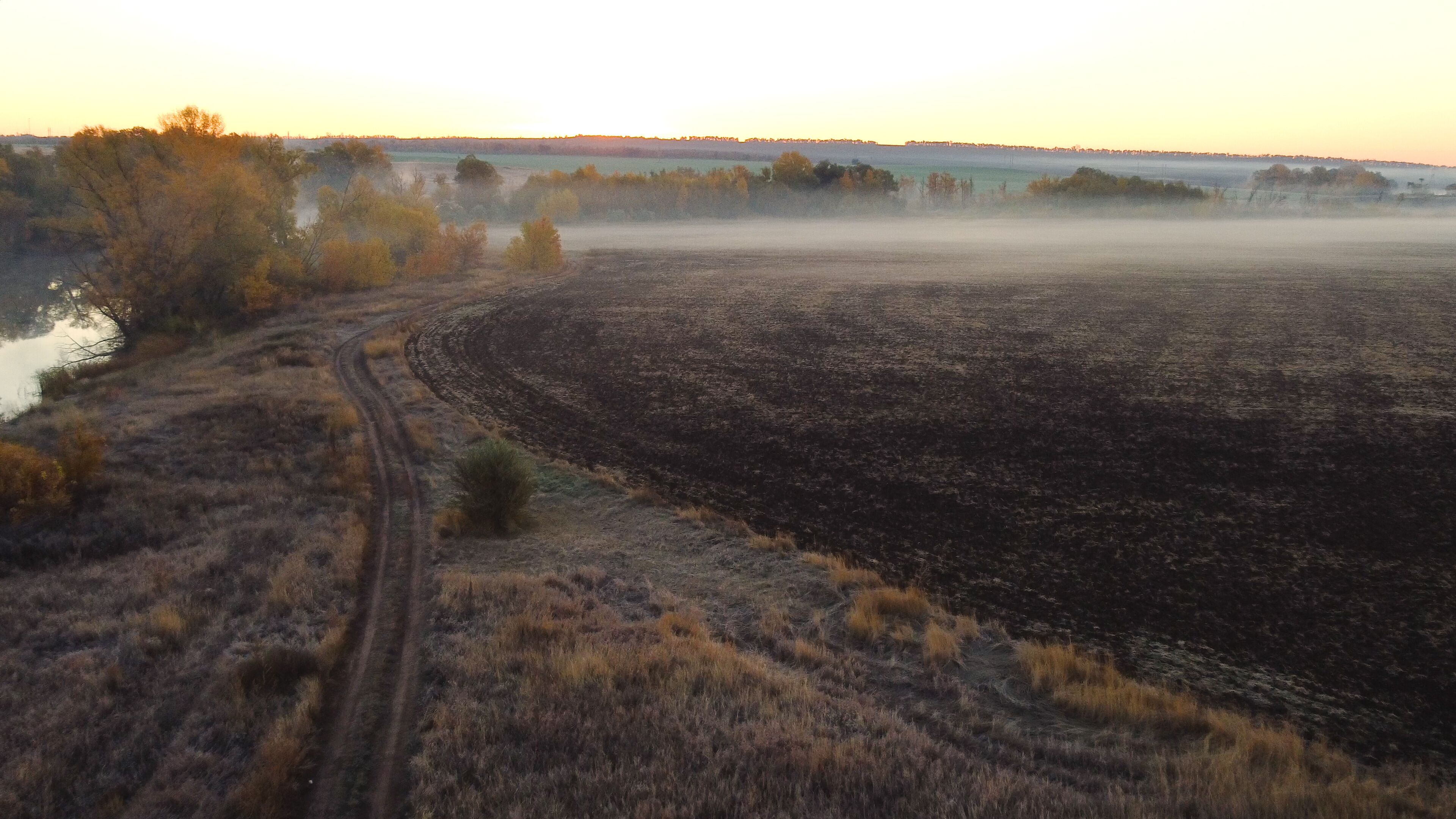 A vast open field with a dirt path leading to a body of water, surrounded by trees and a misty horizon.
