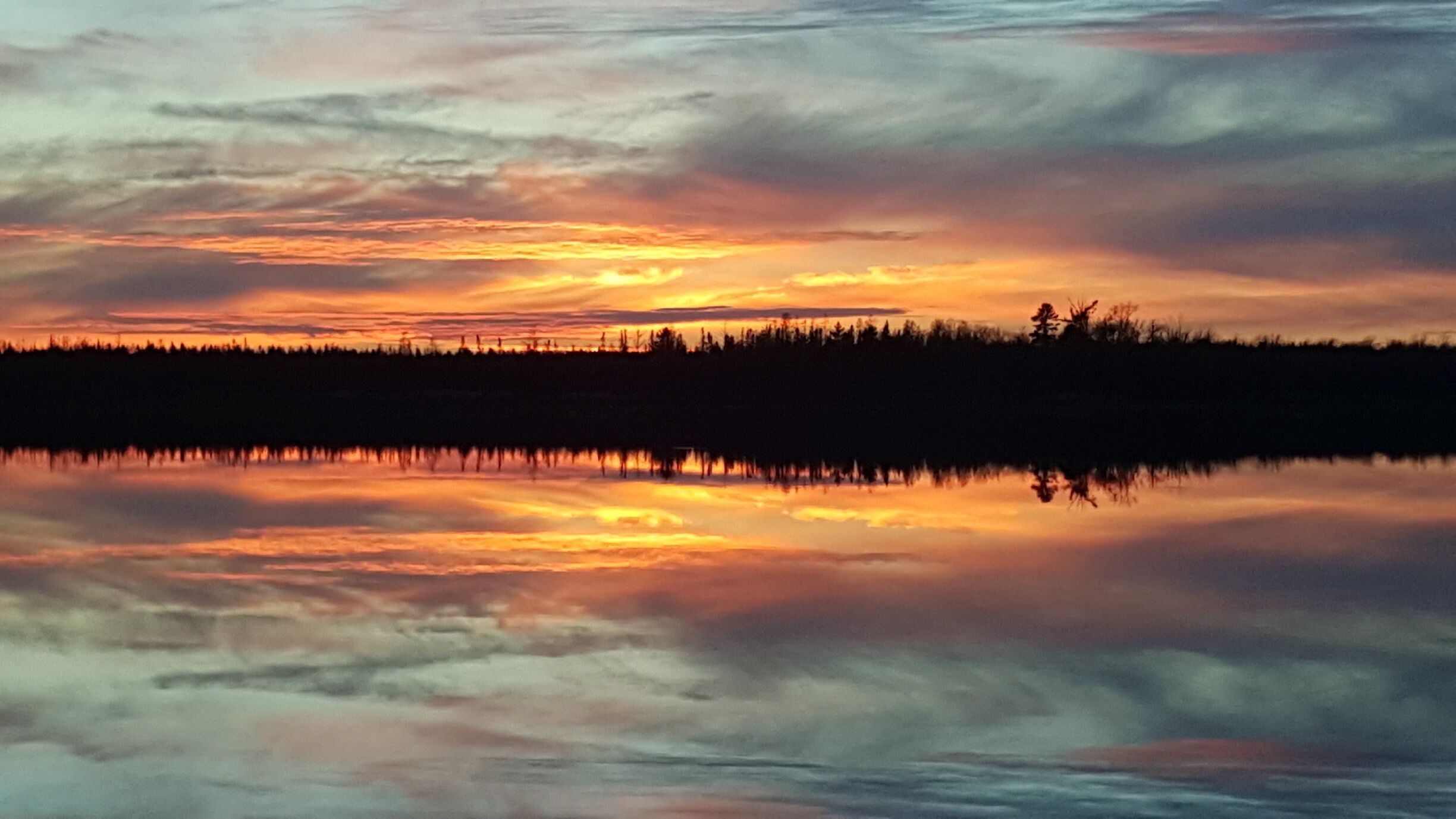 A sunset over a calm lake with a forest silhouette and a cloudy sky.