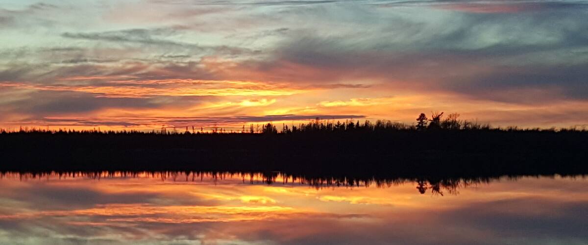 A sunset over a calm lake with a forest silhouette and a cloudy sky.