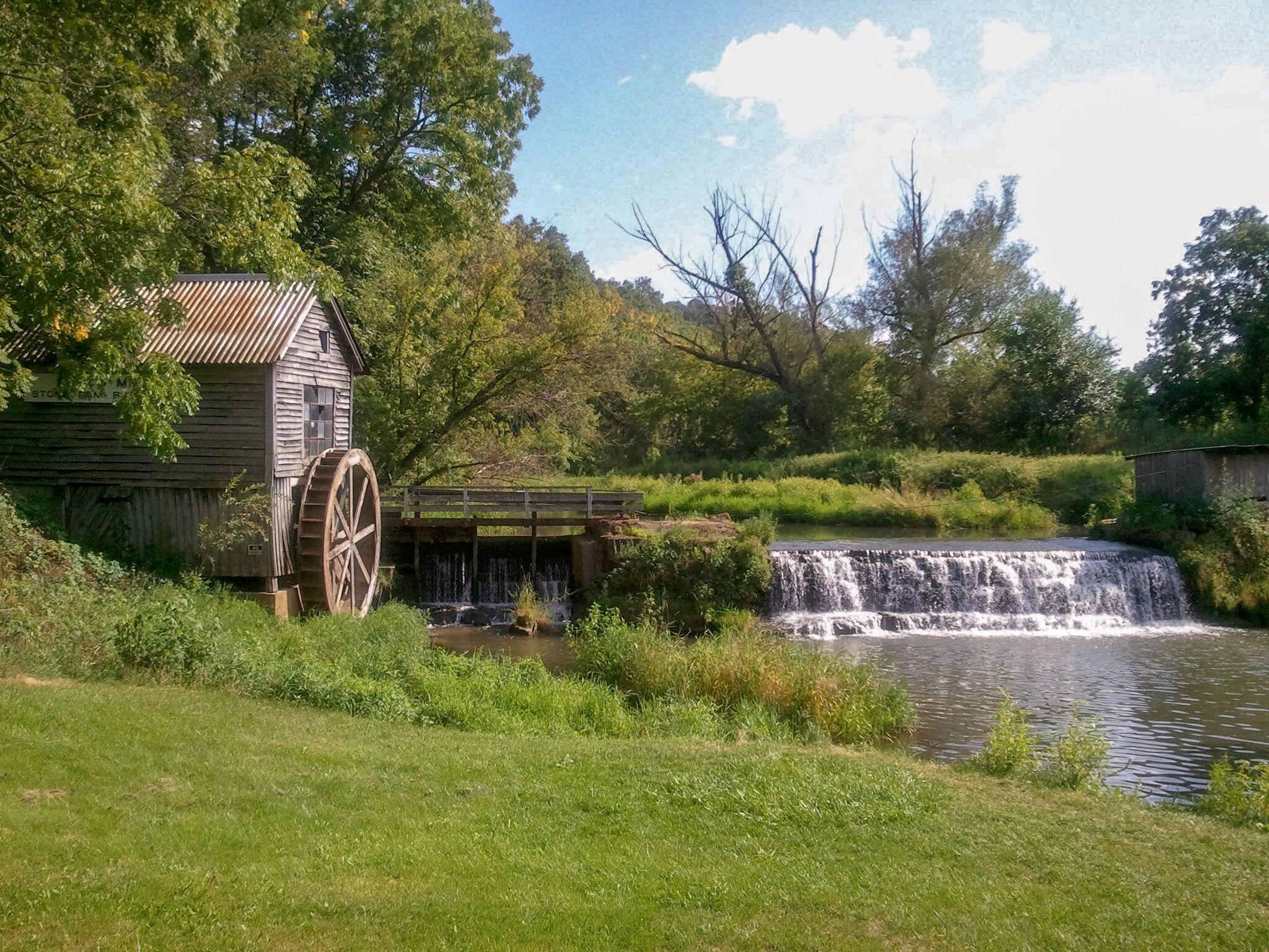 A wooden mill with a large water wheel, a small waterfall, and lush greenery.