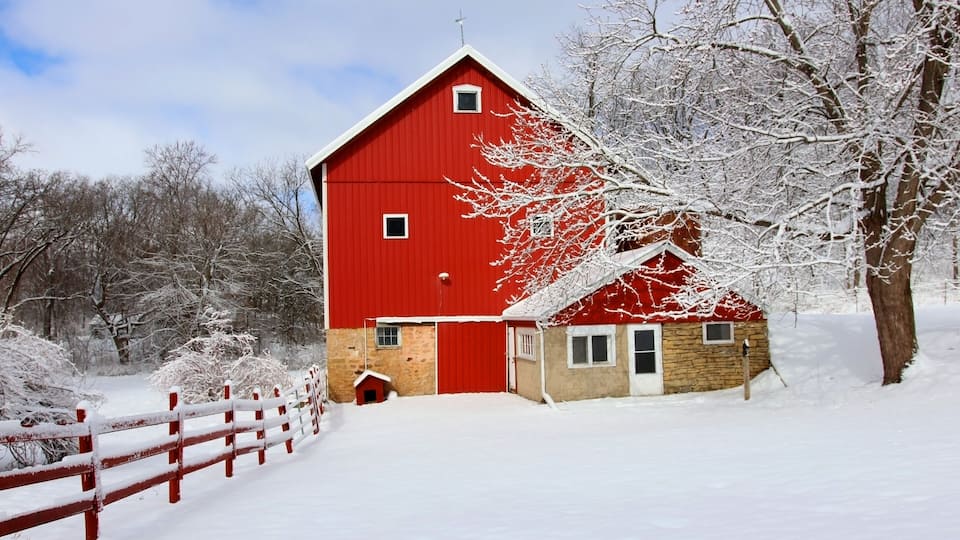Agriculture and rural life at winter background.Rural landscape with red barn, wooden red fence and trees covered by fresh snow in sunlight. Scenic winter view at Wisconsin, Midwest USA, Madison area.